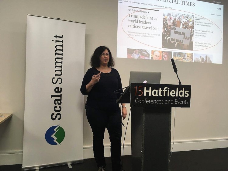 Sarah J Wells stands in front of a lectern with her slides projected on the wall behind her