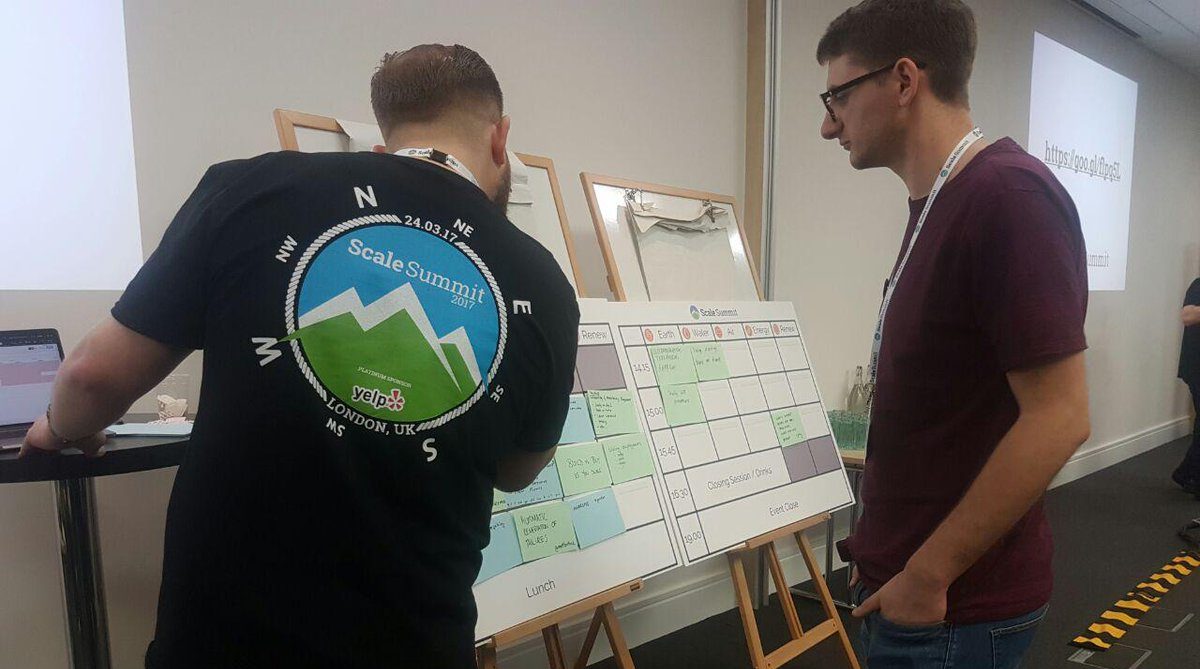 Two people are placing noted index cards on to a whiteboard which has been laid out with day's timings.